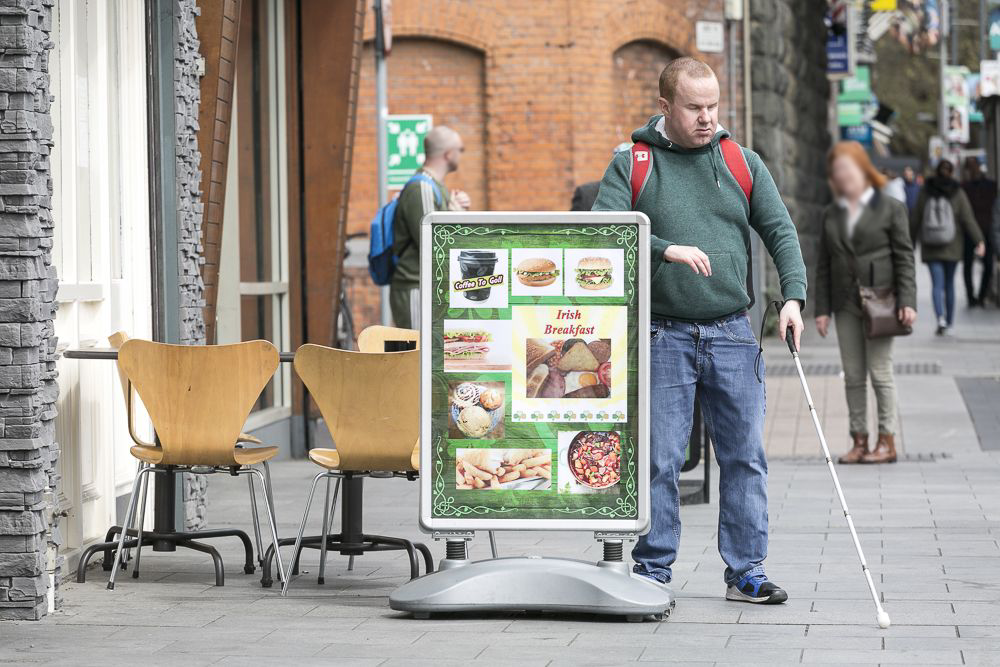 a blind man with his white cane walking on the footpath run into sandwich board