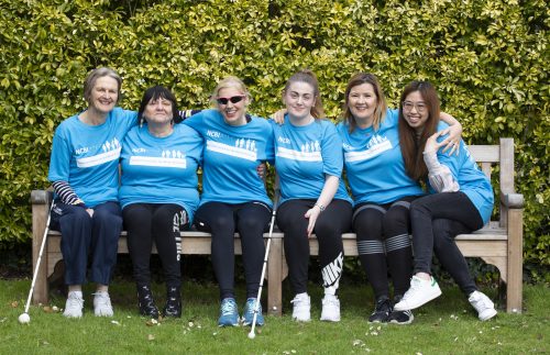 Various Vision Ireland team members in light blue running tops gather around Dr Sinead Kane on a sunny park bench and hold light-hearted stretch poses for the camera.