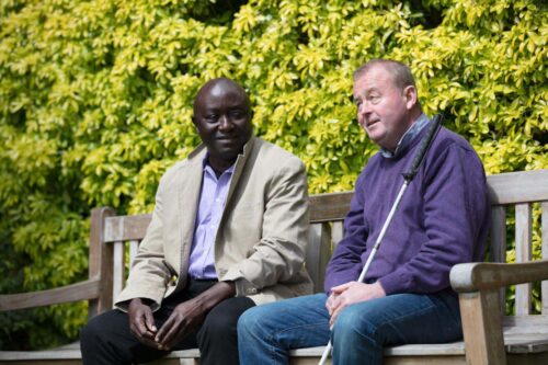 An Vision Ireland service user who has a white cane sitting on a bench alongside another man