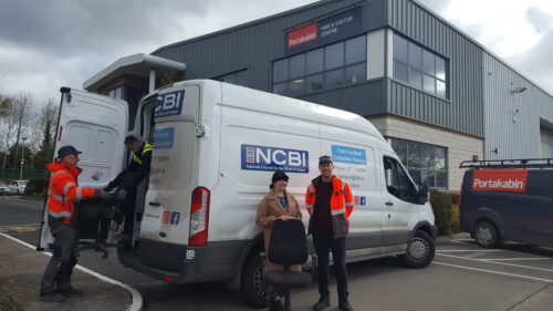 A representative from Portakabin and representatives from Vision Ireland stand in front of a white Vision Ireland van as they accept a donation of furniture from the company, some of which is being loaded into the back of the van