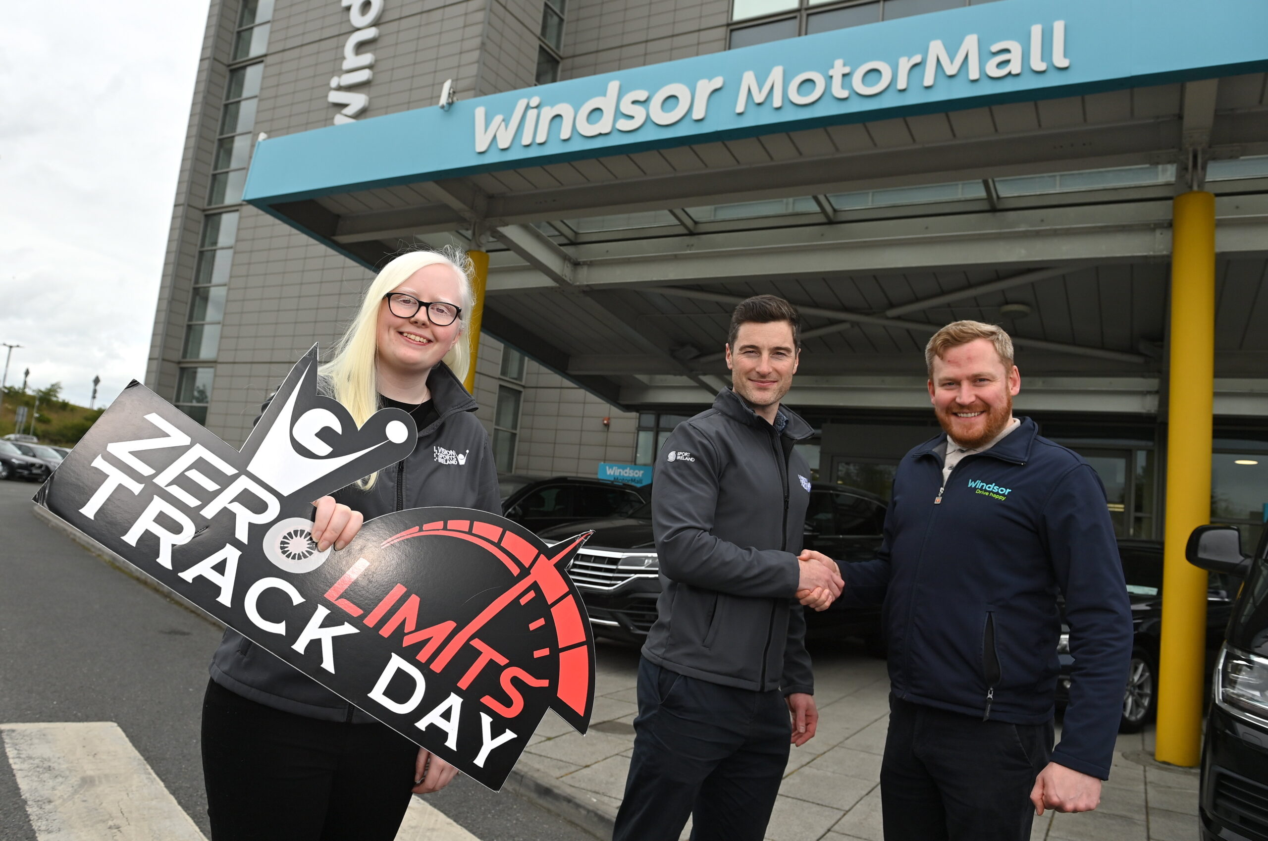 Sara McFadden stands to the left of the picture holding the Zero Limits logo sign. Vision Ireland's Aaron Mullaniff is to the right and he is shaking the hand of a representative from WIndsor Motors.