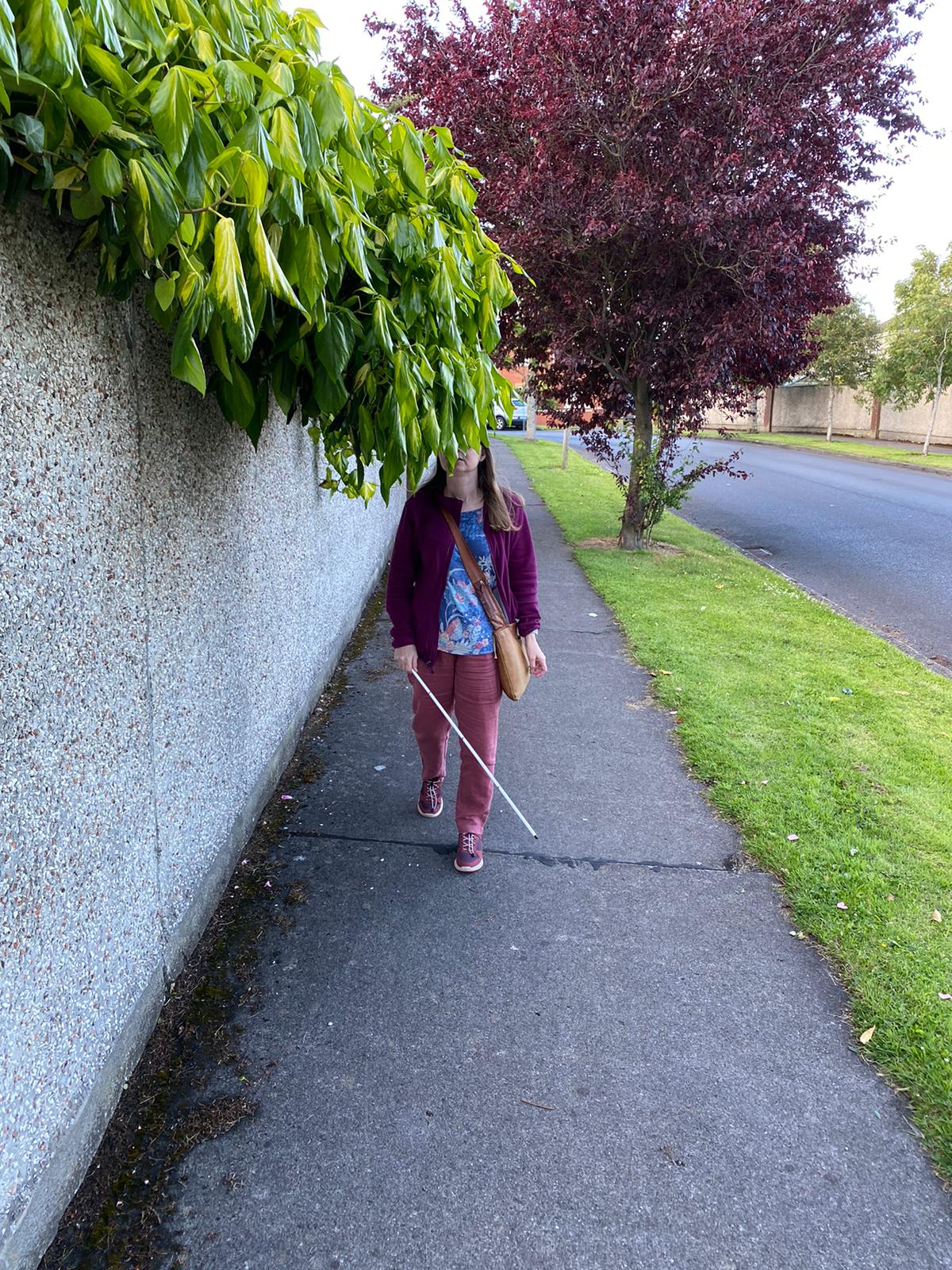 The image shows a woman walking with a white cane into branches which are hanging over a wall along a pathway.