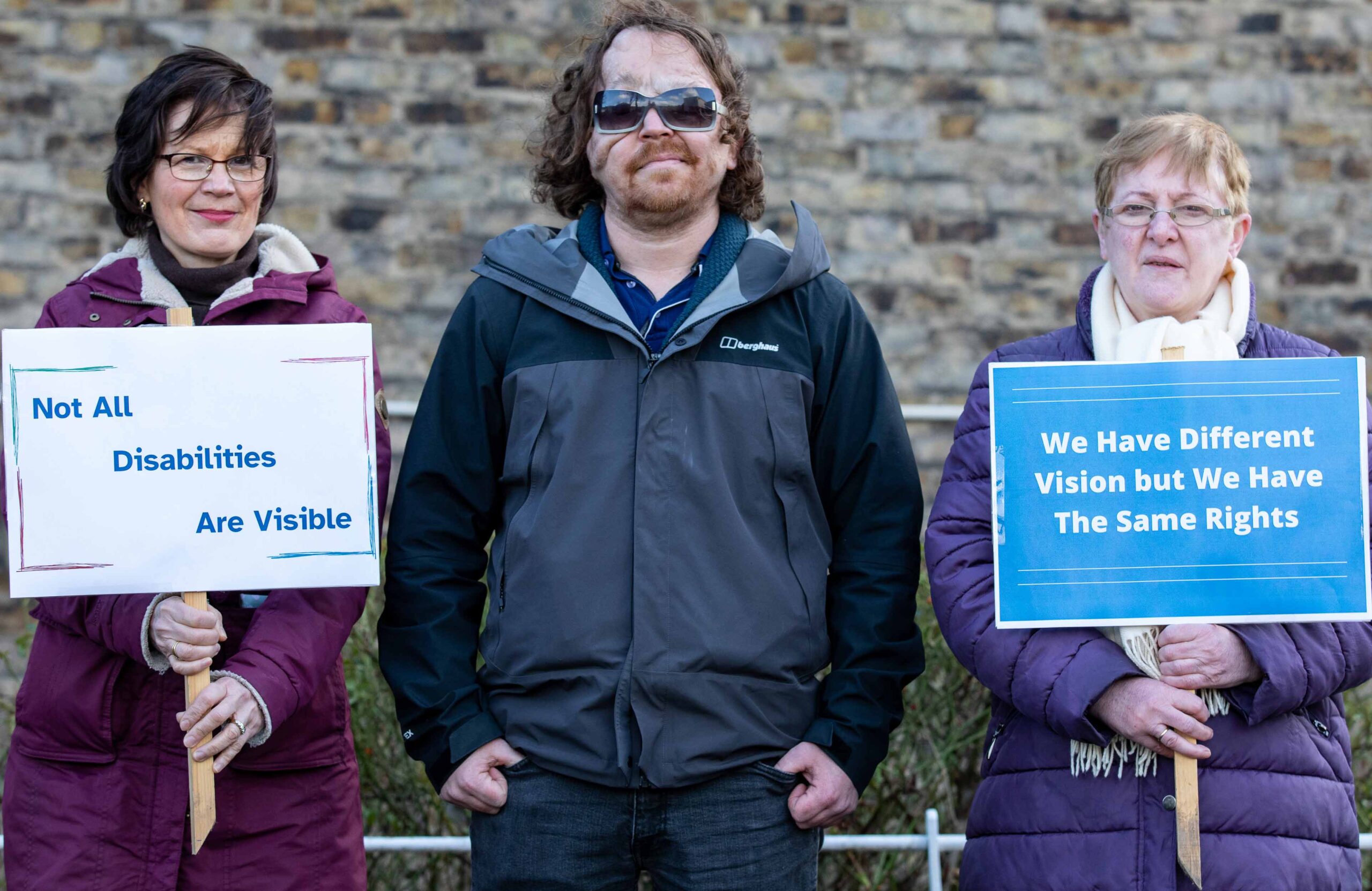 Three Vision Ireland advocates standing together. There is a man in the middle and two women, one either side of him, who are holding signs. The sign on the left reads: Not all disabilities are visible. The sign on the right reads: We have different vision but we have the same rights.
