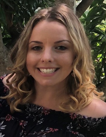 A close up image of Chantelle Smith who is smiling and standing against a background of leaves. Chantelle has blonde, curled hair in the picture.