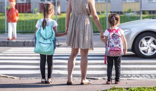 A mother standing at a zebra crossing is holding the hands of her two daughters who are wearing schoolbags.