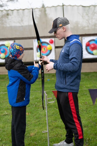 A young boy is holding a bow and arrow and is being directed by a Camp Abilities volunteer to help him shoot at a target