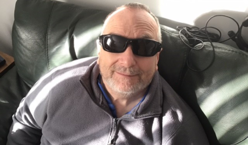 A close up of Gordon McFarlane who is wearing a grey hoodie and he is sitting on a black sofa. Gordon is wearing black sunglasses and is smiling at the camera.