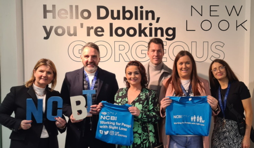 Fiona O'Carroll is standing in the middle of members of Vision Ireland and New Look at the launch of the two organisations' partnership. Everyone in the picture is holding Vision Ireland branding in the form of T-shirts and logos and they're standing against a wall which has a message written on it, which reads: Hello Dublin, you're looking gorgeous.