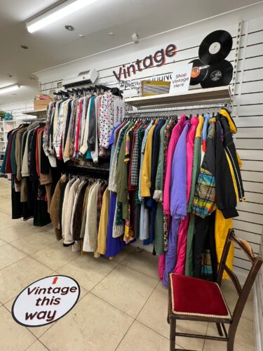 Rails of vintage clothing available in an Vision Ireland store. There is a colourful selection available hanging from the wall, while vintage branding and old-style records decorate the area.
