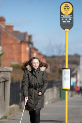 image of a woman who is vision impaired standing at the bus stop with a white cane in her hand
