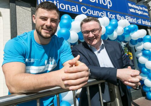 Robbie Henshaw is standing to the left of the image and he is wearing a light blue t-shirt which has Vision Ireland branding on it. Vision Ireland CEO Chris White is standing to the left of Robbie and he is wearing a suit and shirt. Both men are standing in front of an Vision Ireland store which is also decorated with a light blue and white balloon arch.