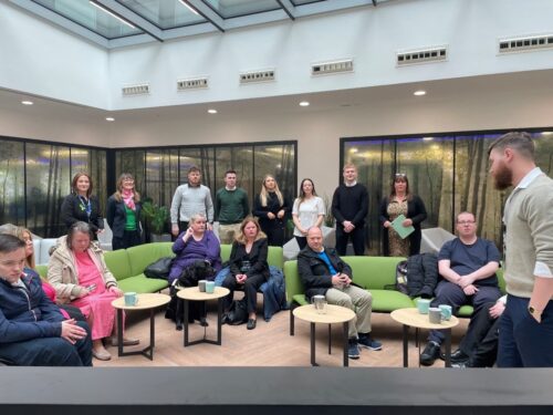 A group of Vision Ireland service users are sitting around in a room on a green sofa as members of the CPL team are also gathered around while a person speaks at the front of the room.