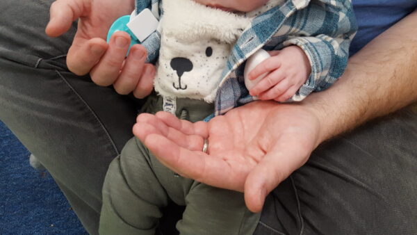 A had has both his hands out for his baby who is sitting on his lap and is also holding a sheep teddy.