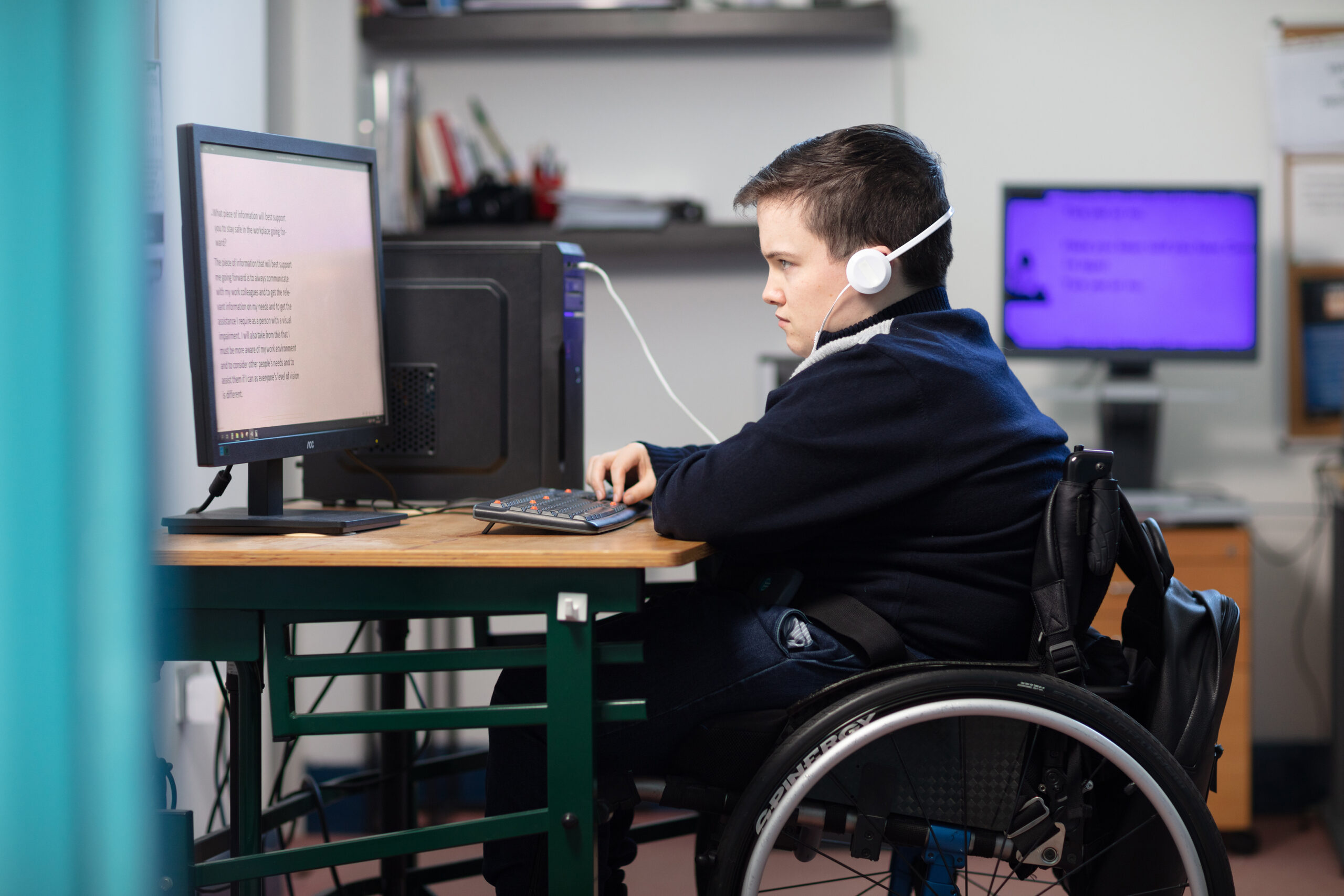 A young man in a wheelchair is sitting at a desk with headphones on and is working on a desktop computer in front of him.