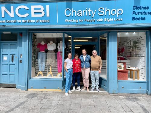 The Vision Ireland Drogheda Team are huddled together and standing in the doorway of the store, which is painted light blue outside and has two windows with a clothing displays.