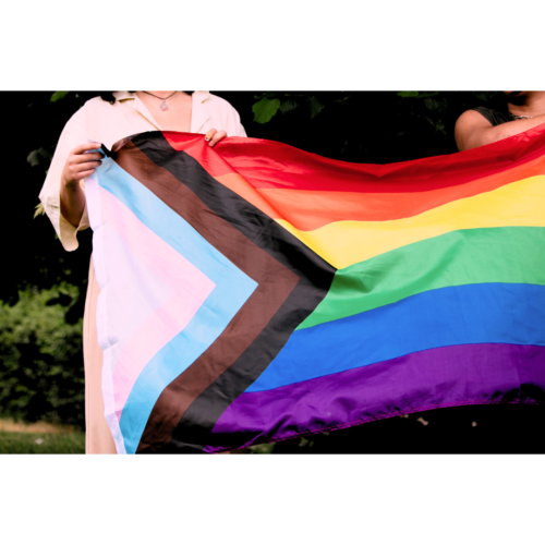 A woman is holding and standing behind a Pride flag which has the red, orange, yellow, green, blue and purple colours stacked one over another, with a triangle design with other strips of colour including black, brown, light blue, pink and white.