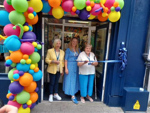 Store manager Jane Dwyer Hall, Carmel McDonnell and Vision Ireland service user Helen Gray cutting the ribbon to open the store, under a multicoloured balloon arch.