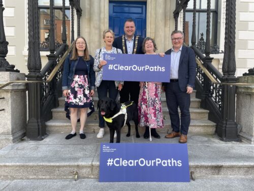 • Madeleine McNamara is standing to the left, alongside Deirdre Deverell with her black guide dog Bruno and beside her is The Lord Mayor of Dublin Daithí de Róiste, Martina Gibney who is standing with her long cane, and Chris White, Vision Ireland CEO. They are holding a blue sign which has white text which reads #ClearOurPaths. An identical sign is also visible on the step below them.