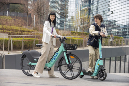 A woman is standing to the left with a Tier e-bike and a man is standing to the right with a Tier e-scooter. Both devices are a light green colour with the Tier logo in blue.