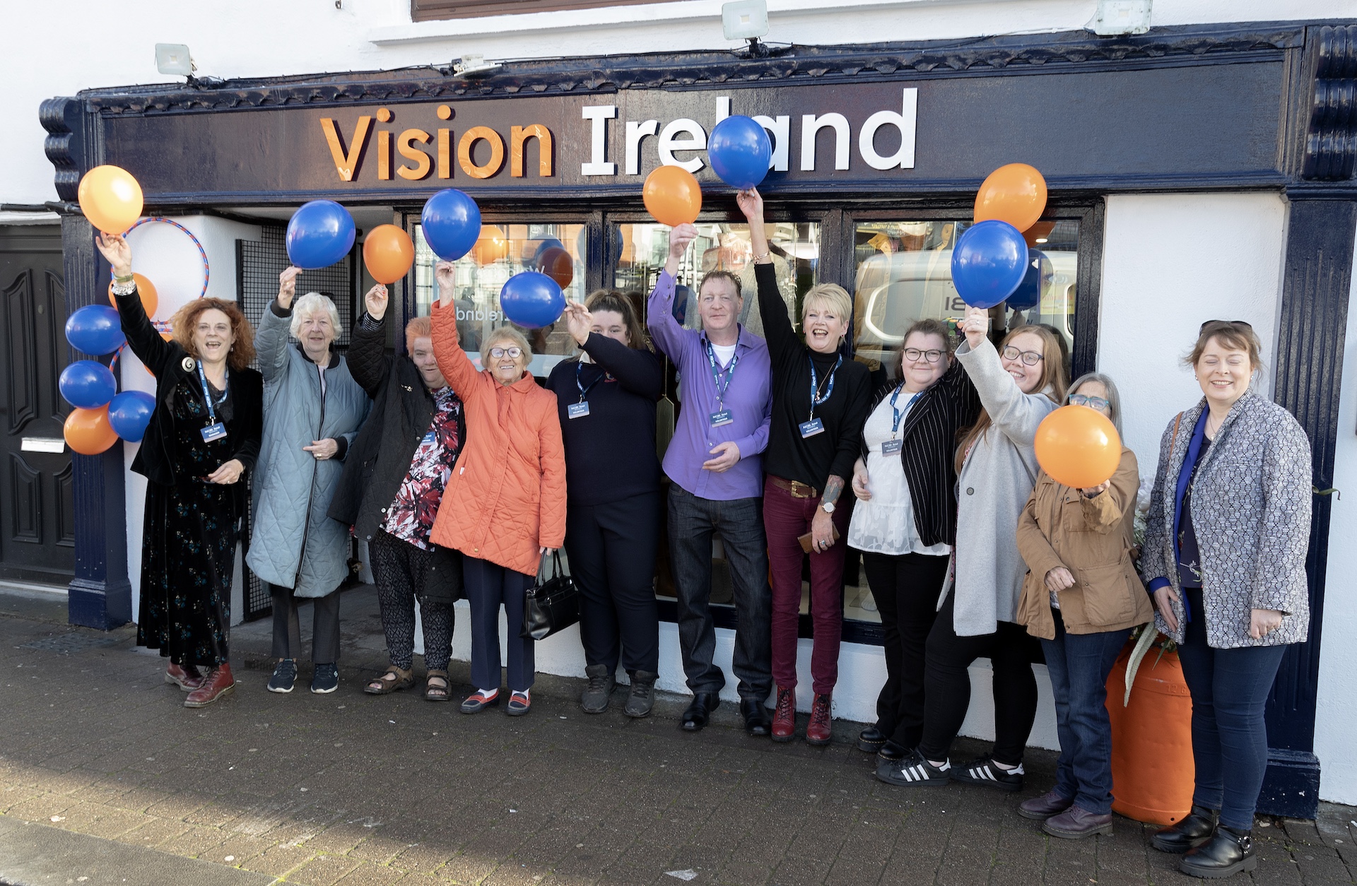 A group of people standing outside a Vision Ireland store and holding up blue, white and orange balloons which match the storefront colours.
