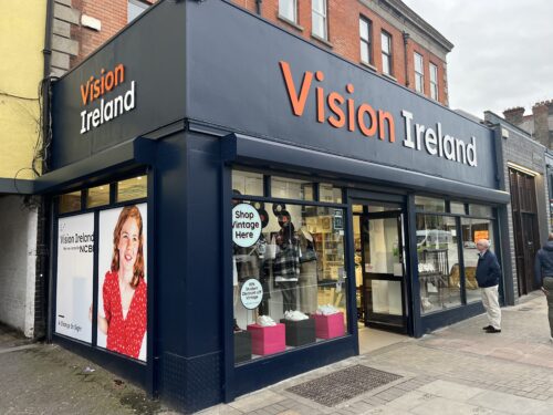 The exterior of the Vision Ireland store on Camden Street. It shows the new orange and white logo over the door and the navy exterior. New Vision Ireland artwork with a young girl with red hair is visible at the side of the store.