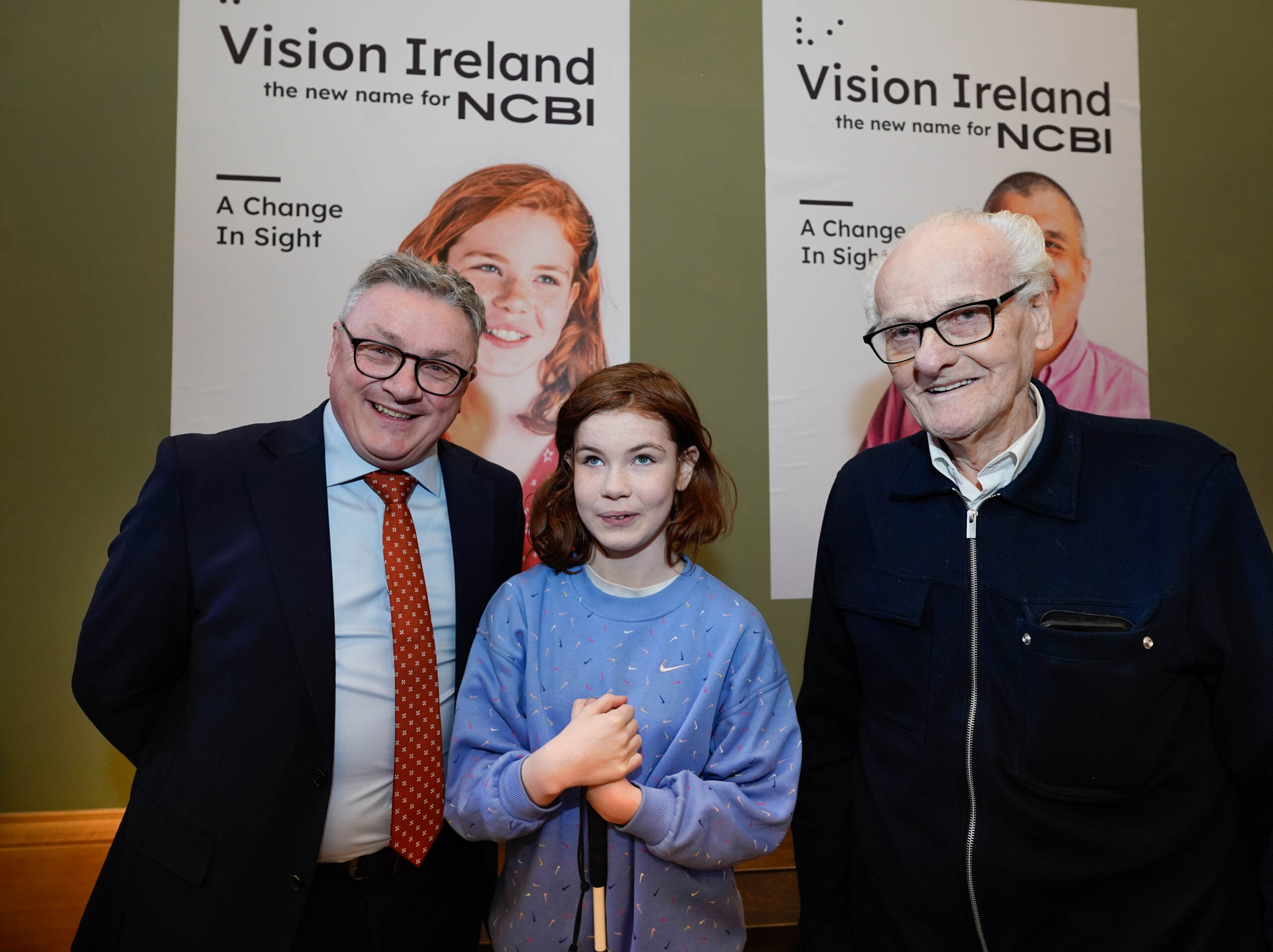 Chris White, CEO of Vision Ireland, is wearing a suit and standing next to Edith Toomey who is holding her white cane and is wearing a blue top, and Lar Keogh who is wearing glasses and a black coat. All three are standing in front of a Vision Ireland poster which features a picture of Edith.
