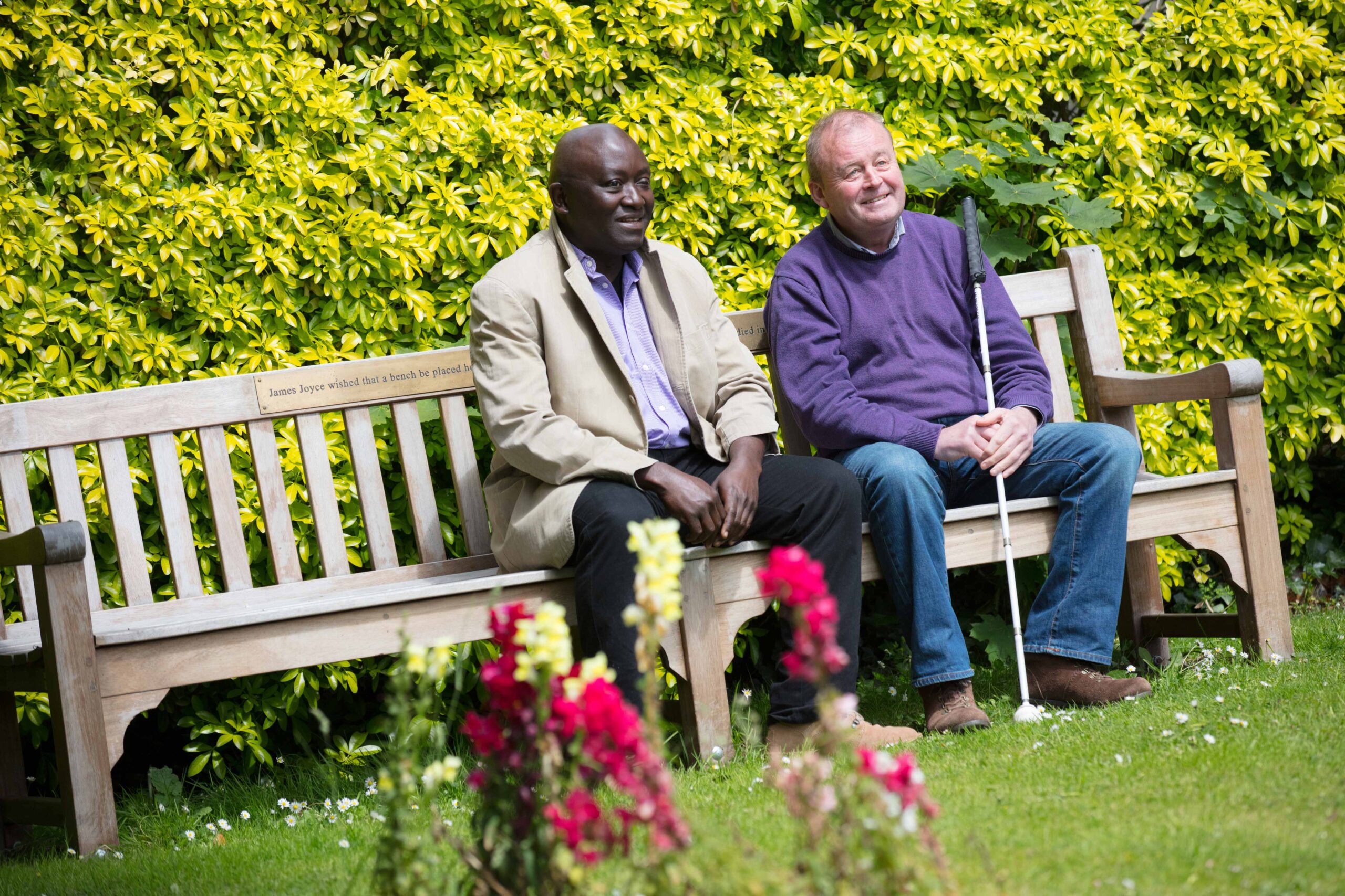 Two men are sitting on a bench in a bright green garden. One of the men is holding a symbol cane and both of them are smiling.