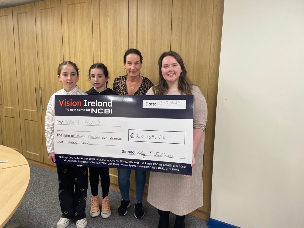 Grace O'Sullivan and her family members pose with a large Vision Ireland cheque to donate following the successful Pedal for Grace event. The cheque is blue and white and has an orange and white Vision Ireland logo on it. The picture is taken in a room in a Vision Ireland office.