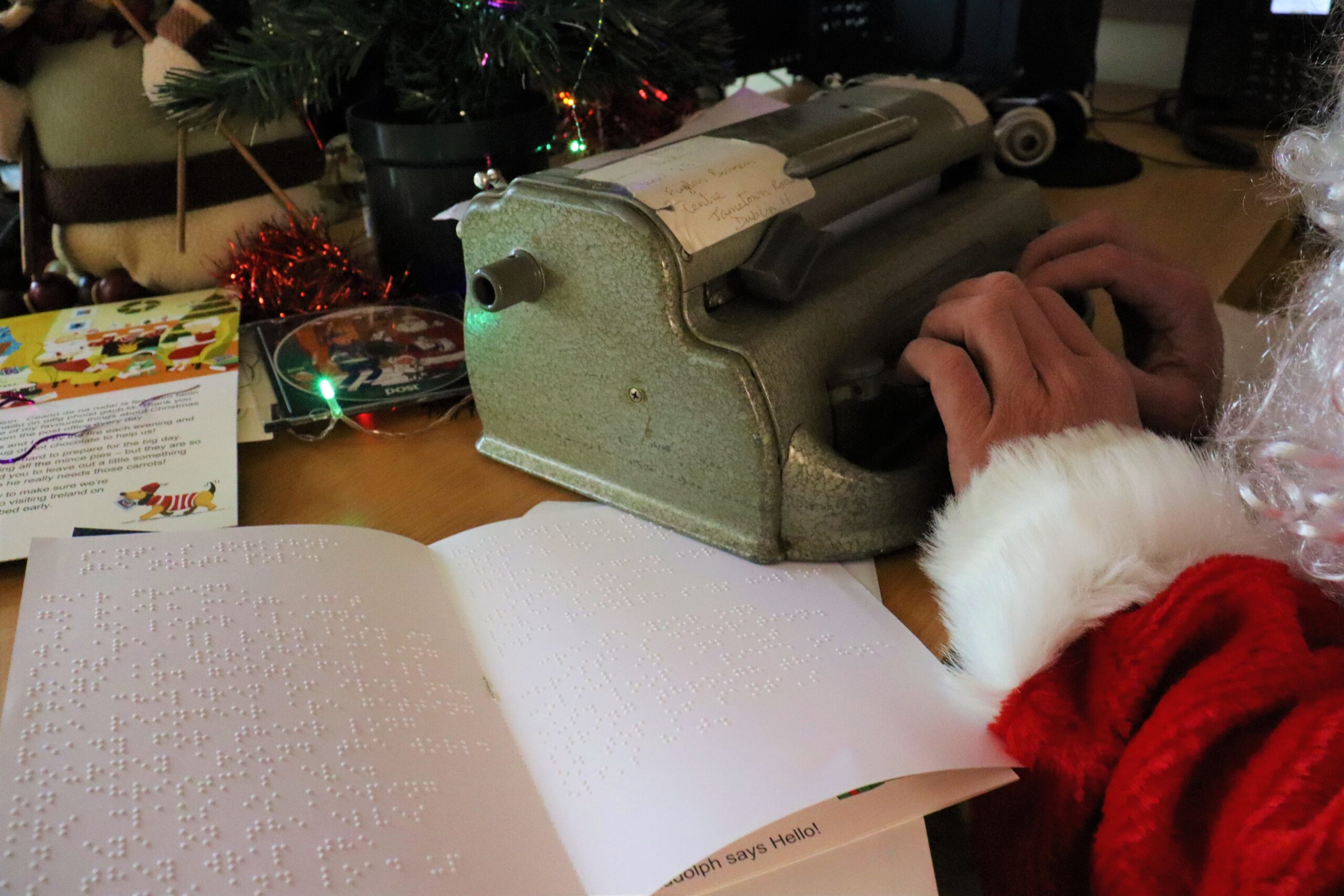 Santa's hands and beard are visible as he types on a Braille machine. He has a Braille letter, an audio CD, and a large print card beside him. He is also sitting beside a Christmas Tree.