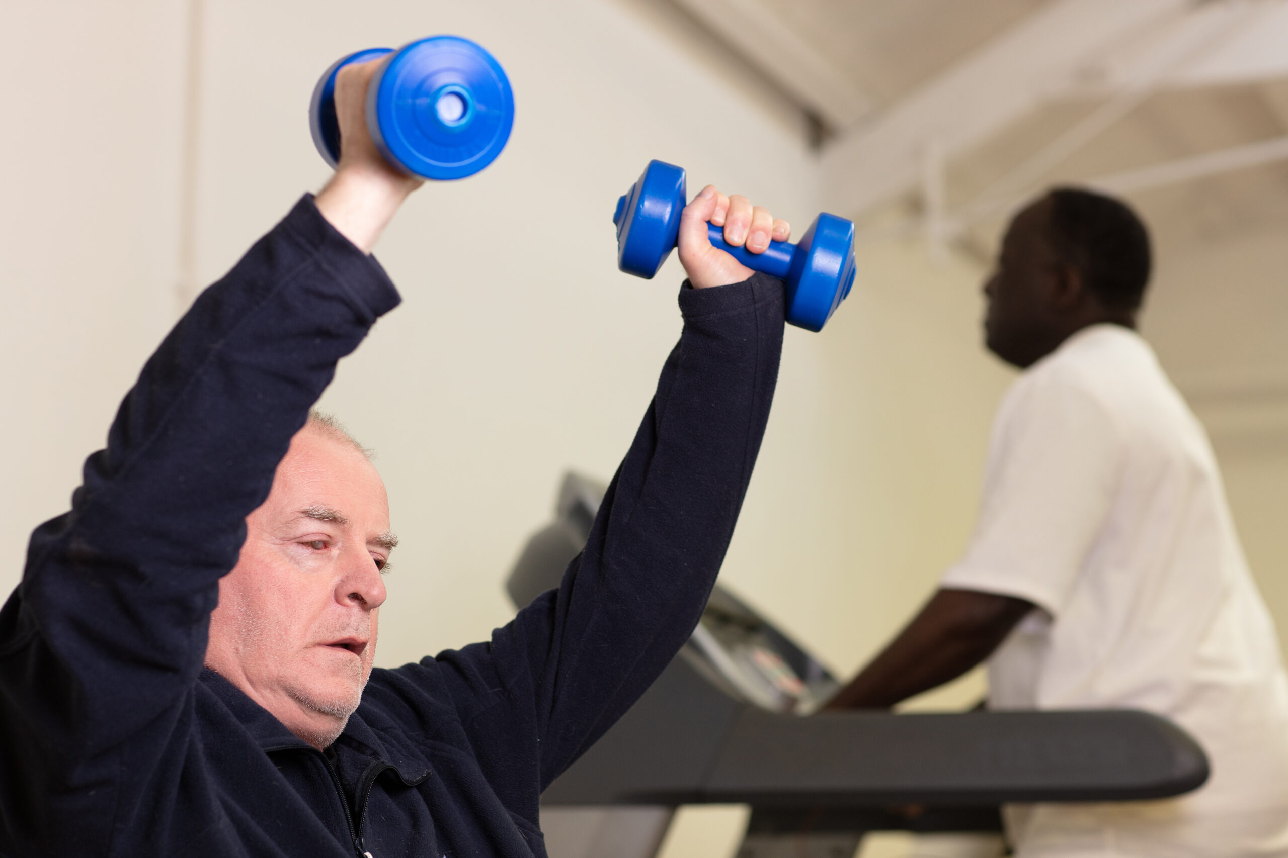 A man is sitting and lifting two blue dumbell weights over his head while another man is blurred in the background running on a treadmill.