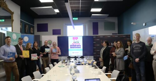 Teams from Vision Ireland, ICBN and St Francis Hospice are gathered together and are standing either side of a long table. They are standing in front of signs promoting each organisation and Bereaved Children's Awareness Week. Some team members are holding versions of the books which have been made accessible.