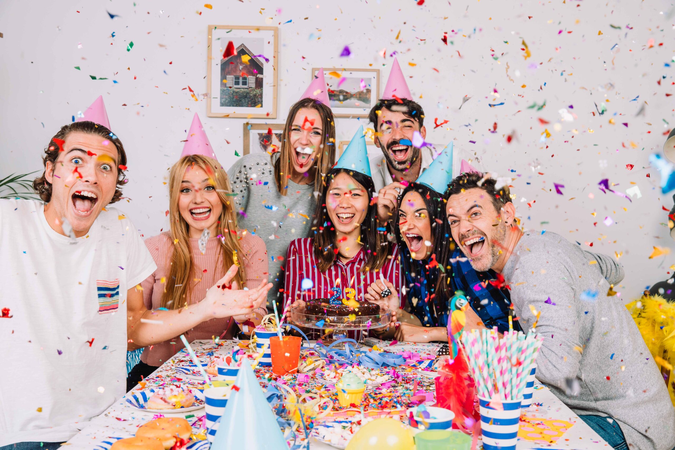 A group of young men and women are sitting around a table which has a birthday cake and decorations. There is multicoloured confetti floating in the air.