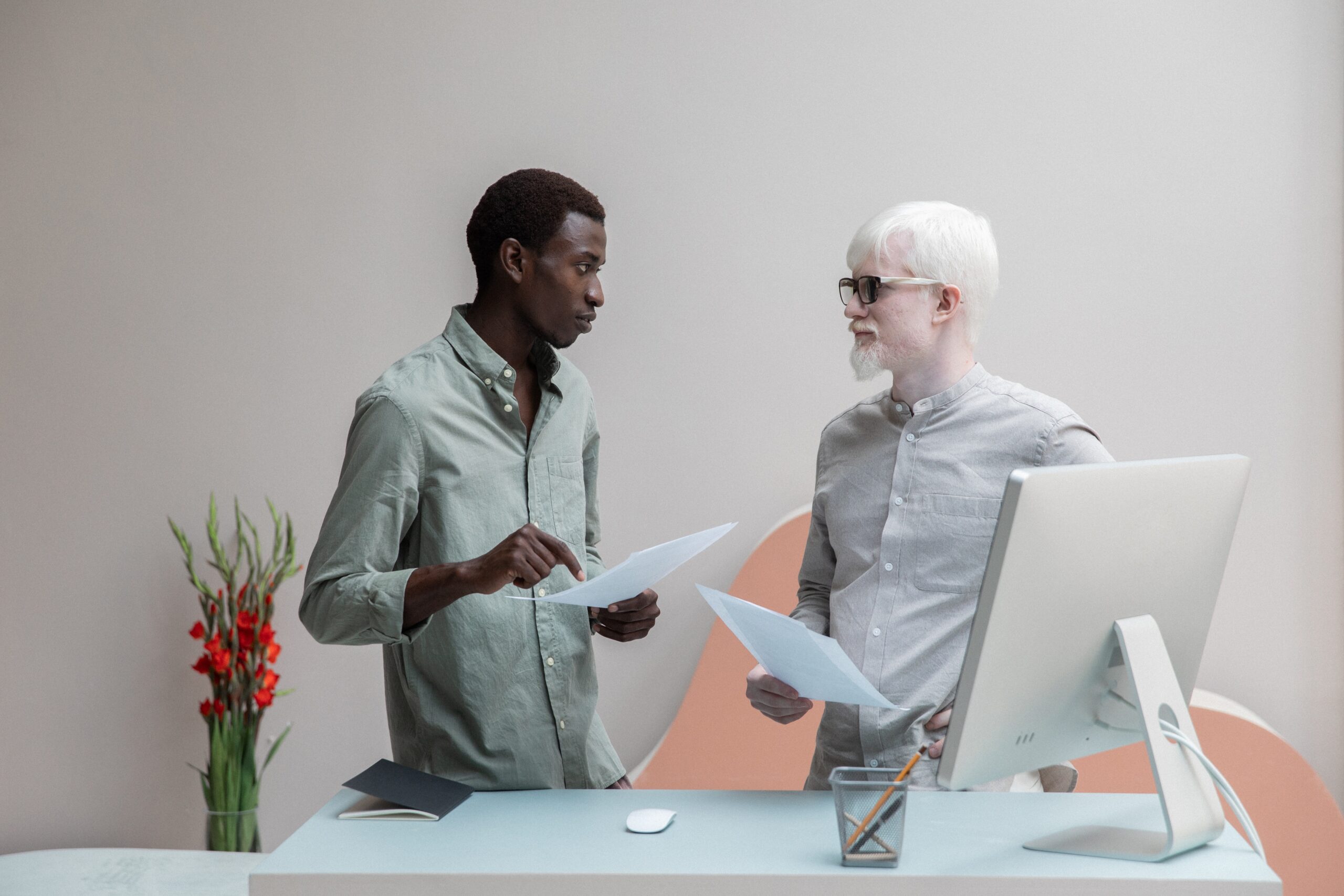 : Two people standing next to each other in an office setting. There is a white desk with a computer in front of them, and they are both holding some papers while they seem to be in a conversation.